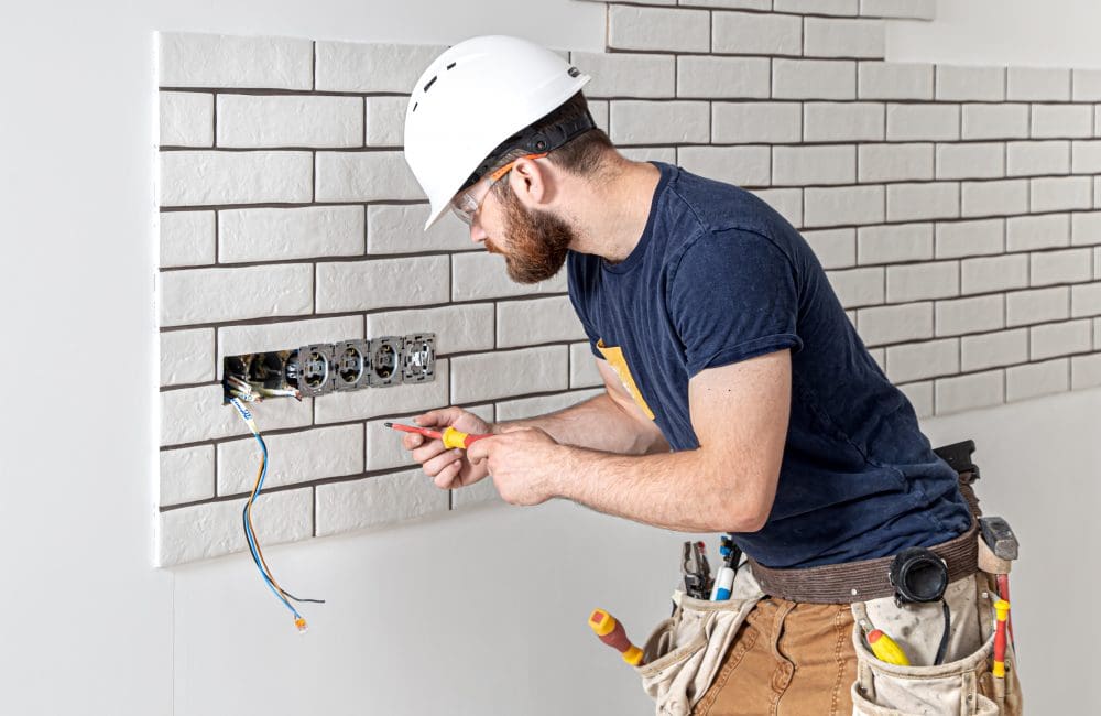 Electrician builder in a white helmet at work, installation of sockets, and switches. Electrician construction worker with a beard in overalls during the installation of sockets. Home renovation concept.